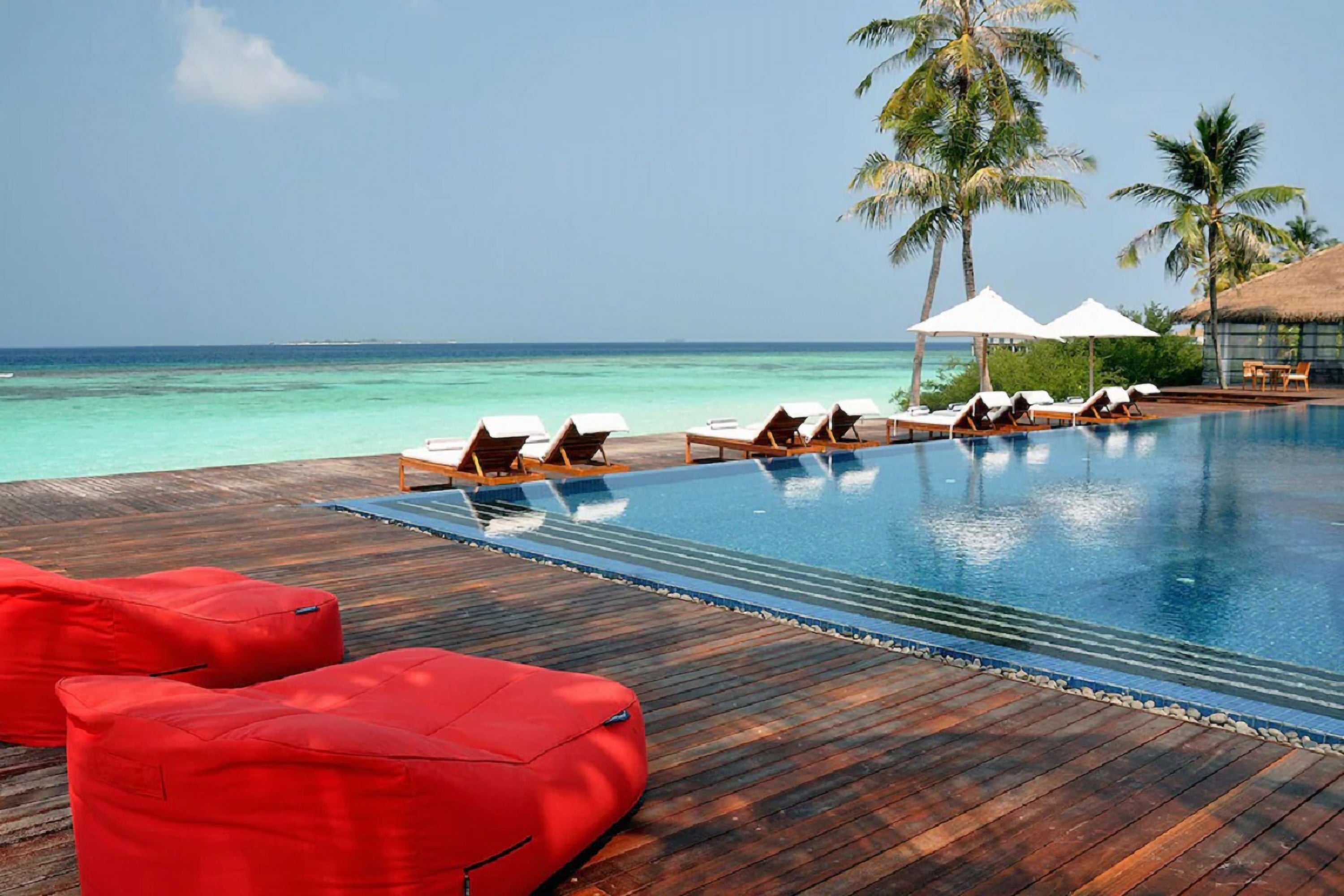 Noku Maldives Resort pool area with Satellite Twin Sofa bean bags, wooden deck, and palm trees by the ocean.